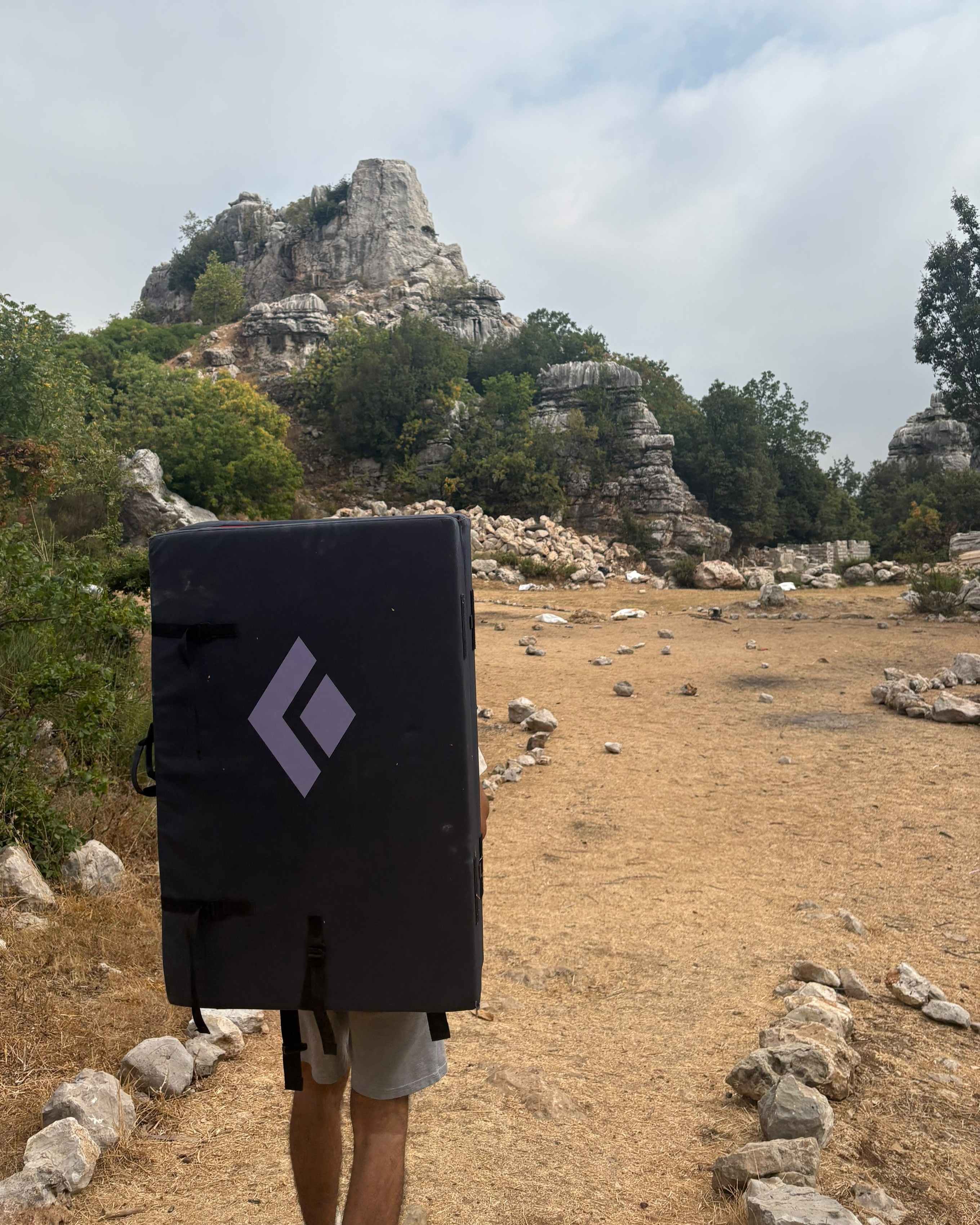 A climber carrying a crash pad beneath a rocky crag.